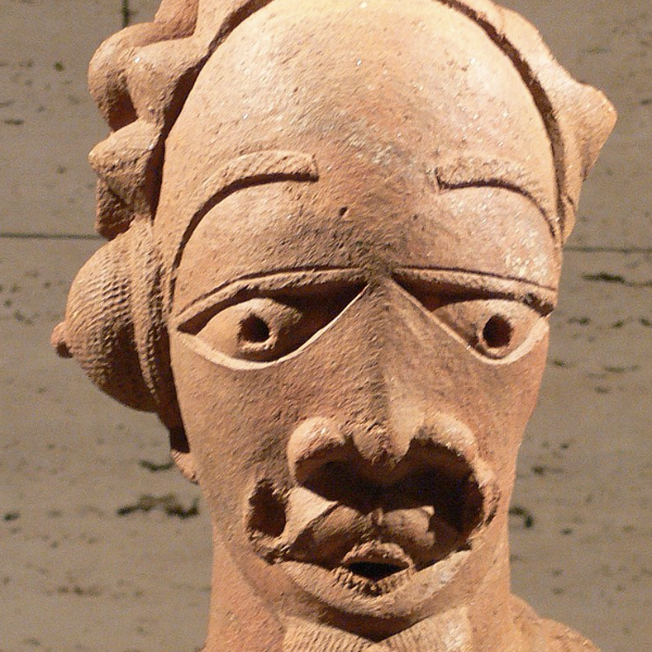 A terracotta sculpture of a male figure with defined facial features, a hairstyle with multiple buns and caps over the ears, and numerous necklaces and other jewelry.