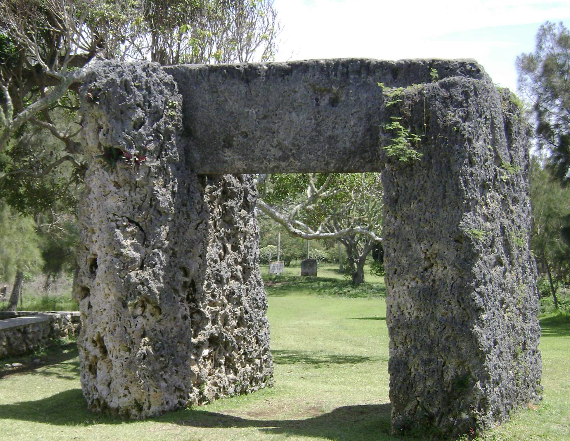 A stone monument with two stones acting as posts and a third stone sitting vertically atop the others. The monument sits on grass and there are trees seen in the background. 