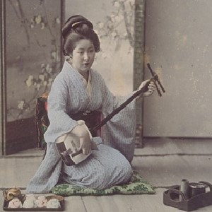 A woman dressed in a kimono and hair in Japanese style sits in a room playing a shamisen. A tray with a tea set and a tobacco tray are placed next to her, and a screen is placed at her back.