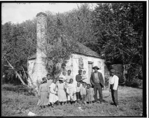 Black and white photograph of what appears to be a black family from what appears to be the antebellum period, with several children and a few adult men and women, standing in front of a white house with a chimney. Behind the house is a wooded area.
