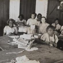 Black and white photo of 10 girls and one teacher seated at tables with needles, fabric, and sewing machines.